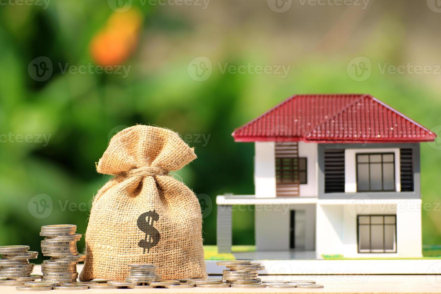 Burlap bag with dollar symbol next to stacks of coins and miniature house photo