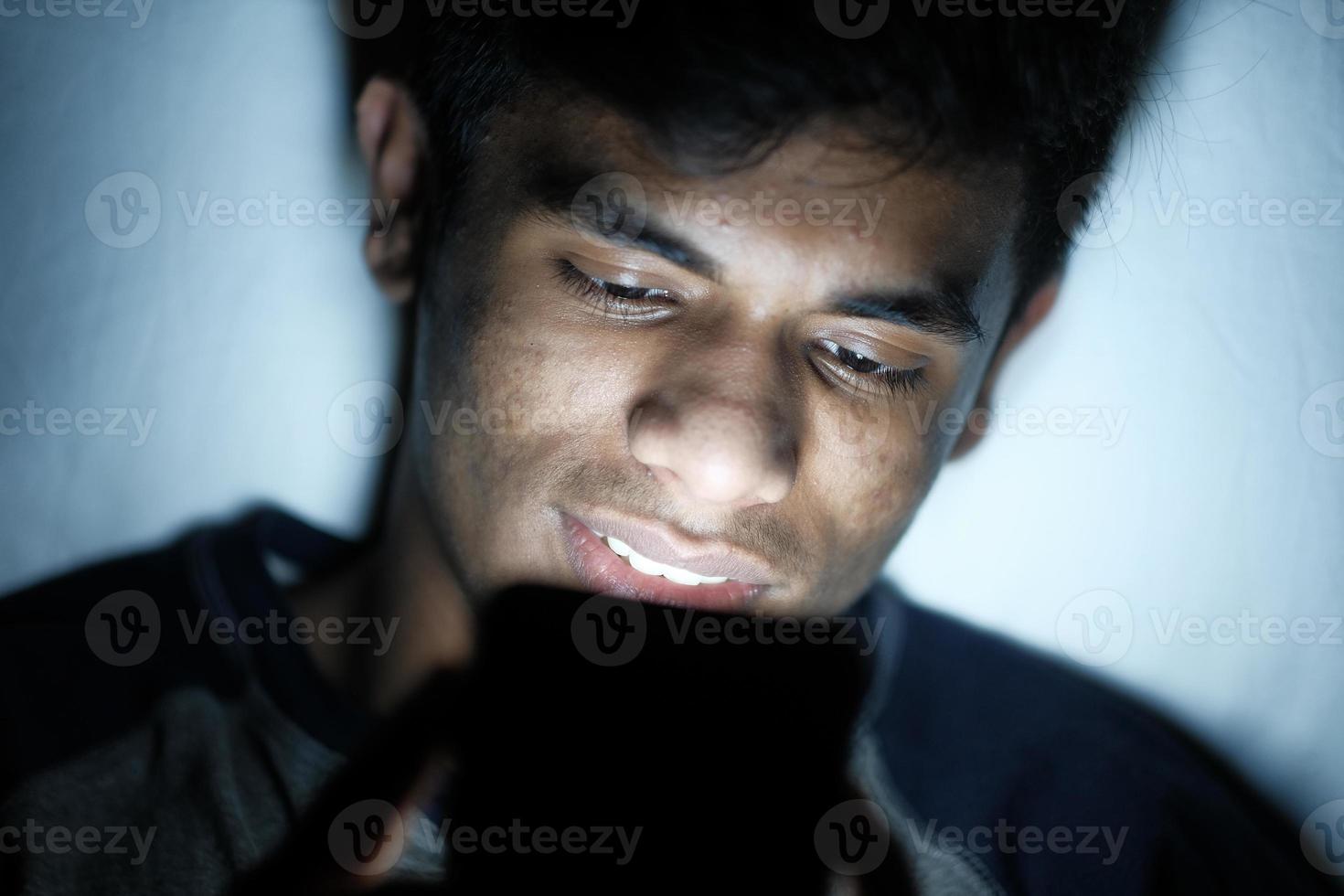 Young man sitting on bed using smartphone at night photo