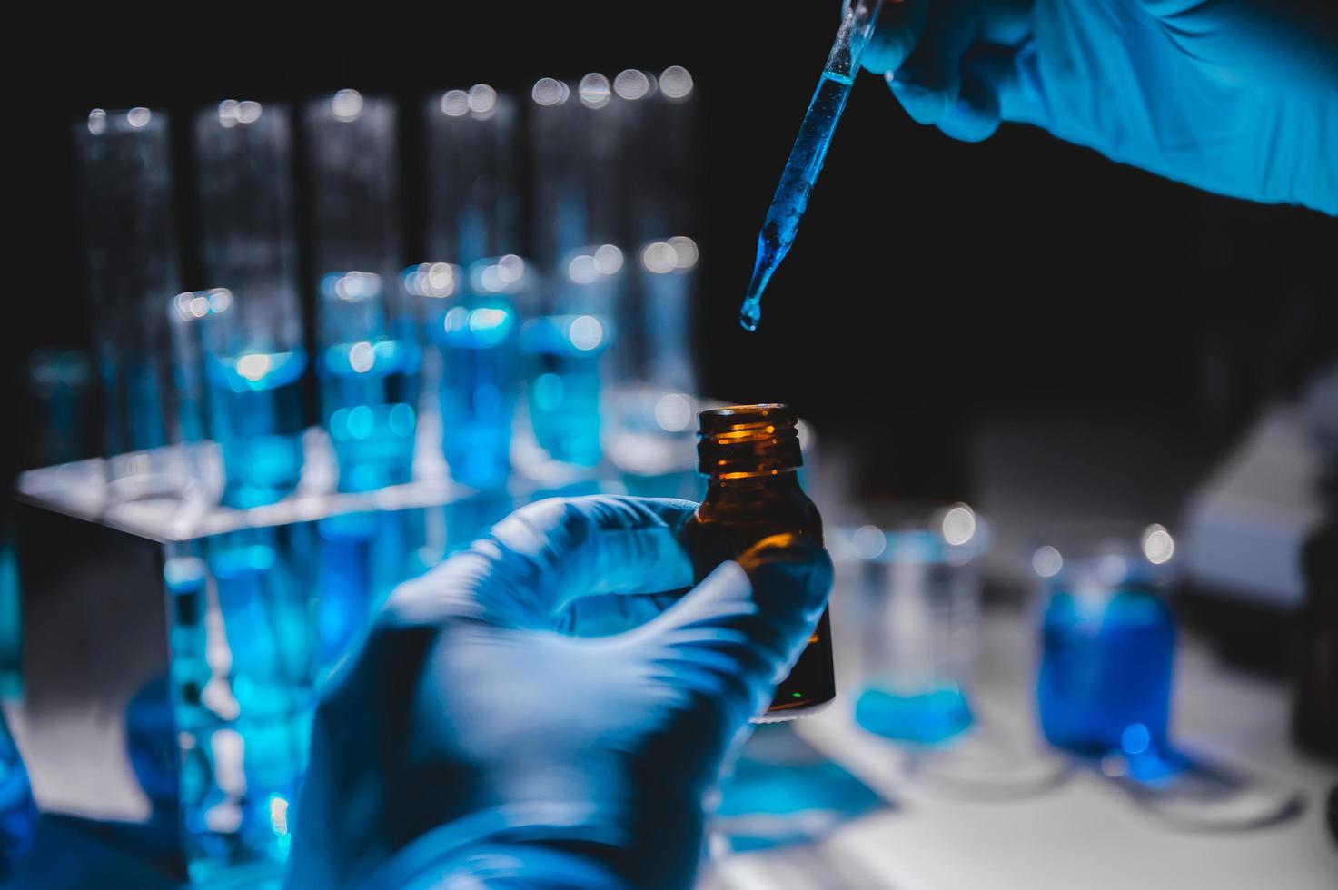 Hands in blue gloves using dropper to put blue liquid into a vial with vials of blue liquid in the background photo