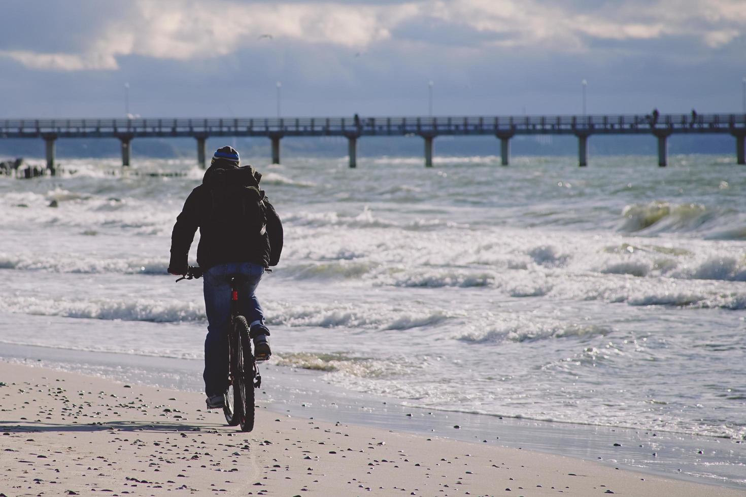 Ciclista en ropa oscura paseos en una playa frente al muelle con nublado cielo azul en zelenogradsk, Rusia foto