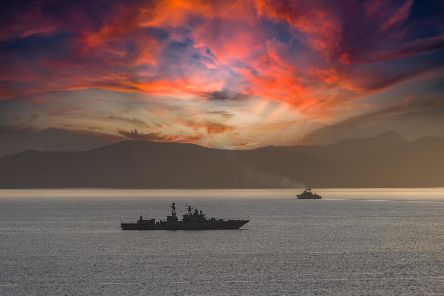 Seascape with silhouetted warships in water next to mountains at sunset photo