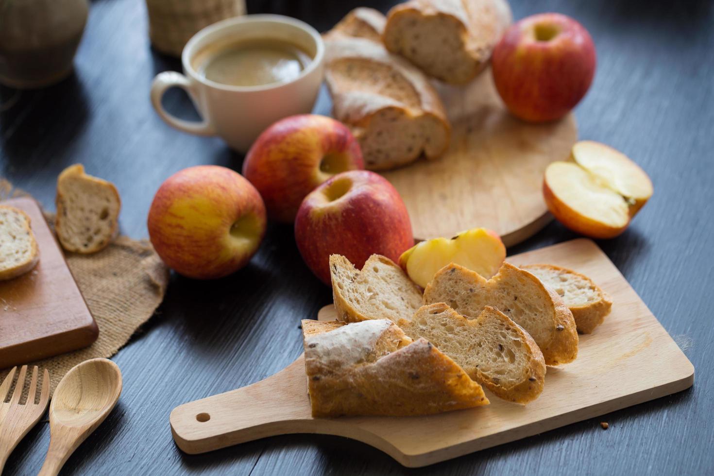 Manzanas enteras y en rodajas con baguette en rodajas sobre tabla de madera con utensilios de madera y una taza de café en una mesa de madera oscura. foto