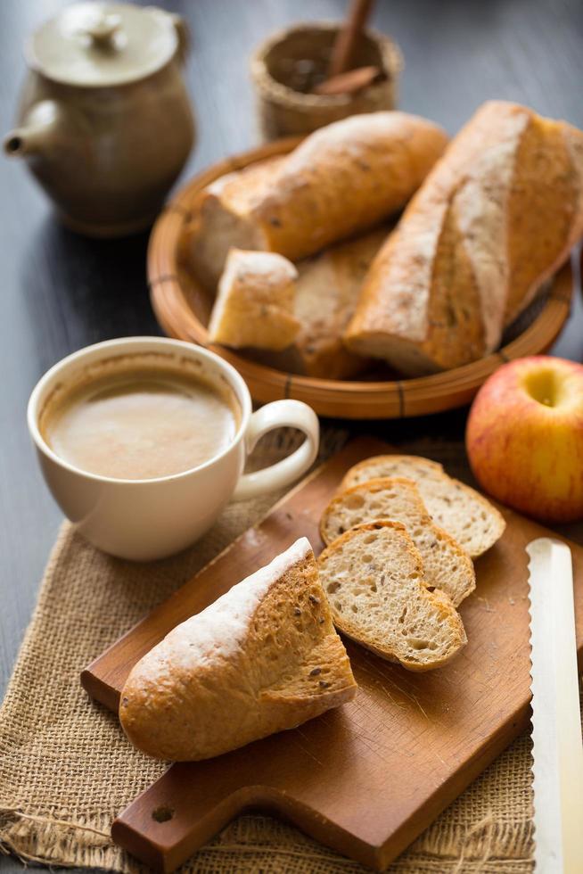 Whole and sliced apples with sliced baguette on wooden board with a cup of coffee and bread knife on a dark wooden table photo