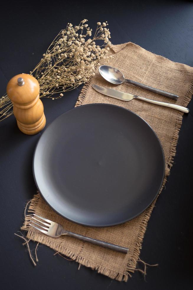 Vista superior de la mesa vacía de plato negro, tenedor, cuchillo, cuchara y molinillo de pimienta en arpillera sobre una mesa negra foto