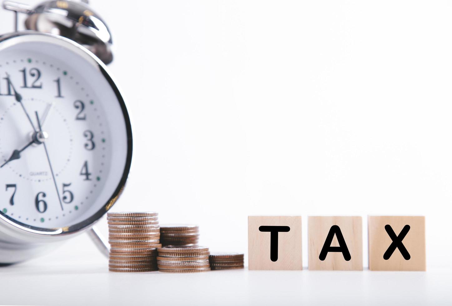 Time to pay tax clock, stacked coins, and wooden block on white background. Taxation and Annual tax concept photo