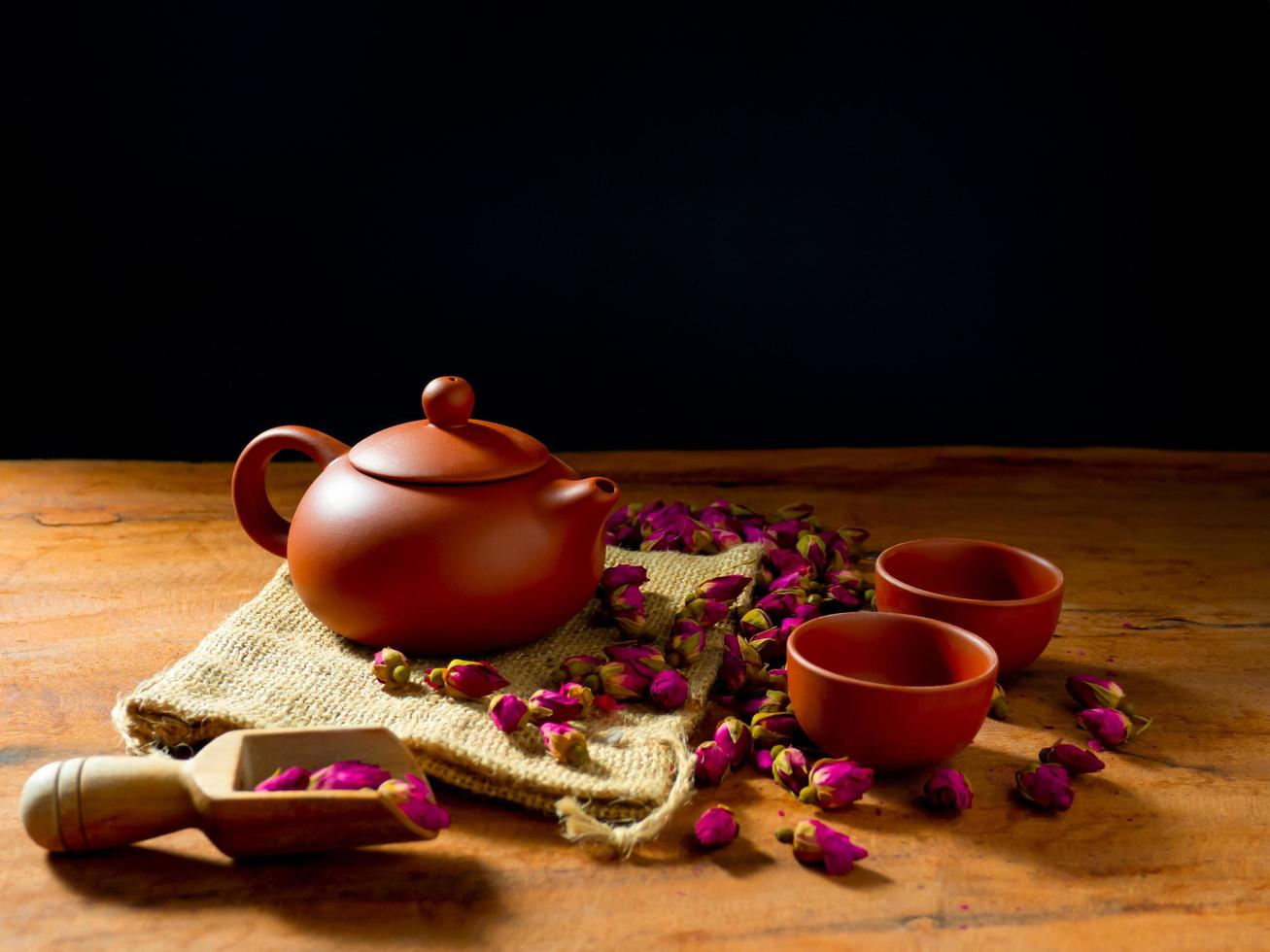 Teapot and teacup with rose tea leaves on wooden table and black background photo