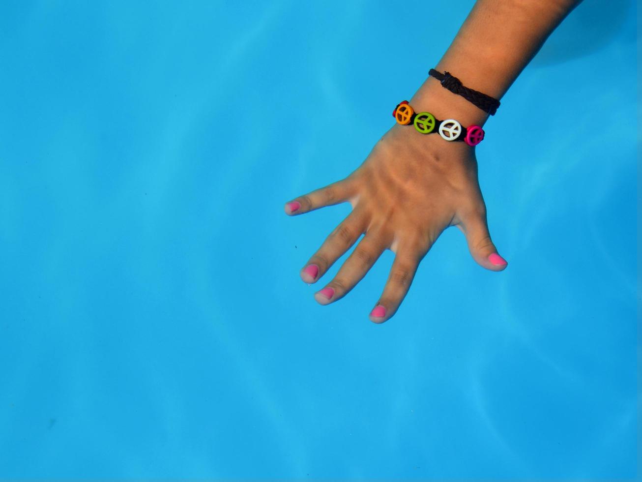 una mano con una pulsera y uñas de colores bajo el agua azul de una piscina en el verano foto