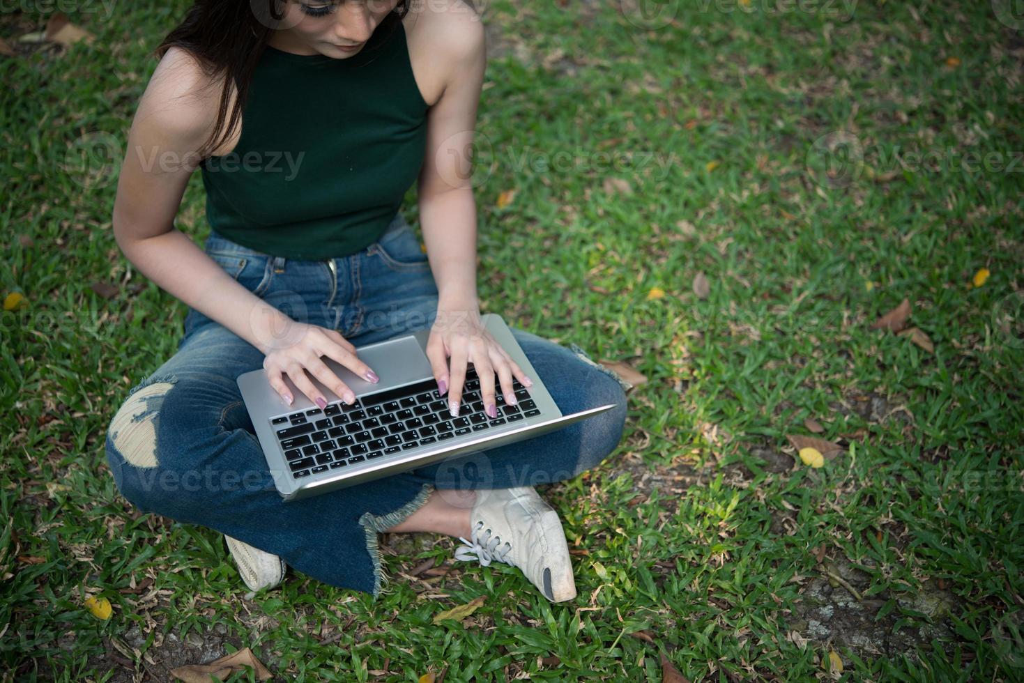 Hermosa mujer joven sentada sobre la hierba verde y usando la computadora portátil en el parque foto