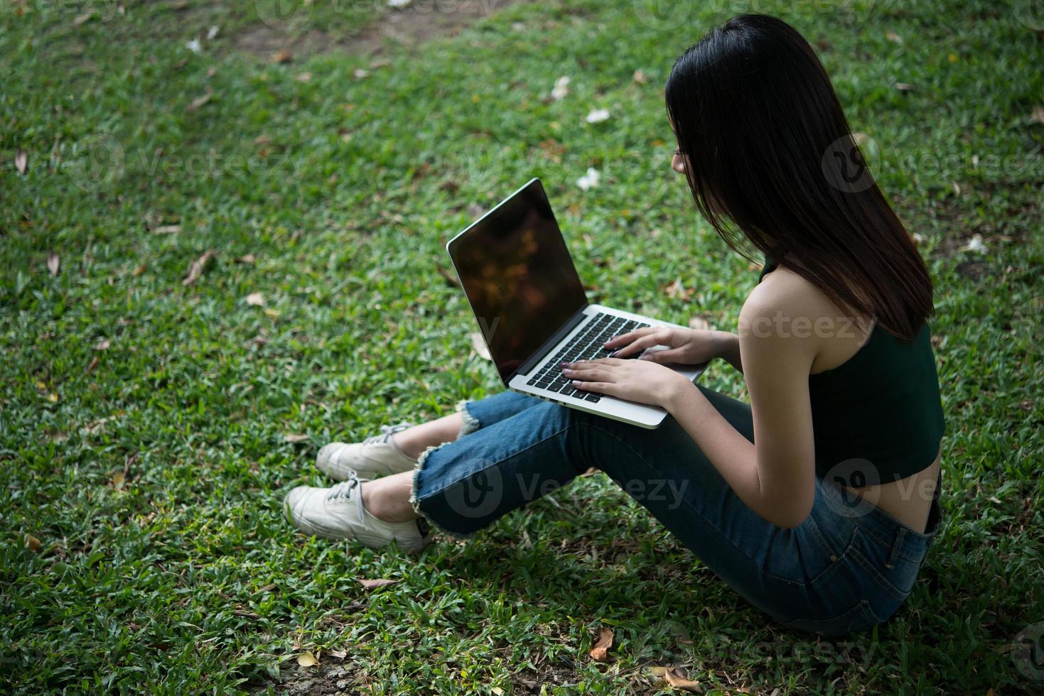 Young beautiful woman sitting on green grass and using laptop in the park photo