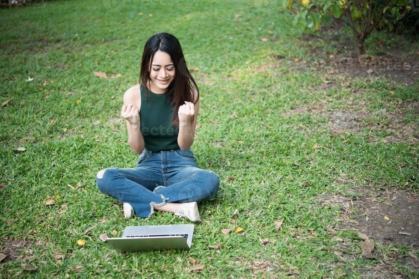 Hermosa mujer joven sentada sobre la hierba verde y usando la computadora portátil en el parque foto