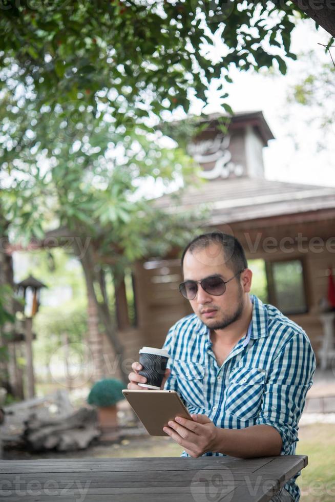 El joven se sienta al aire libre en una mesa de madera y se relaja con una tableta foto