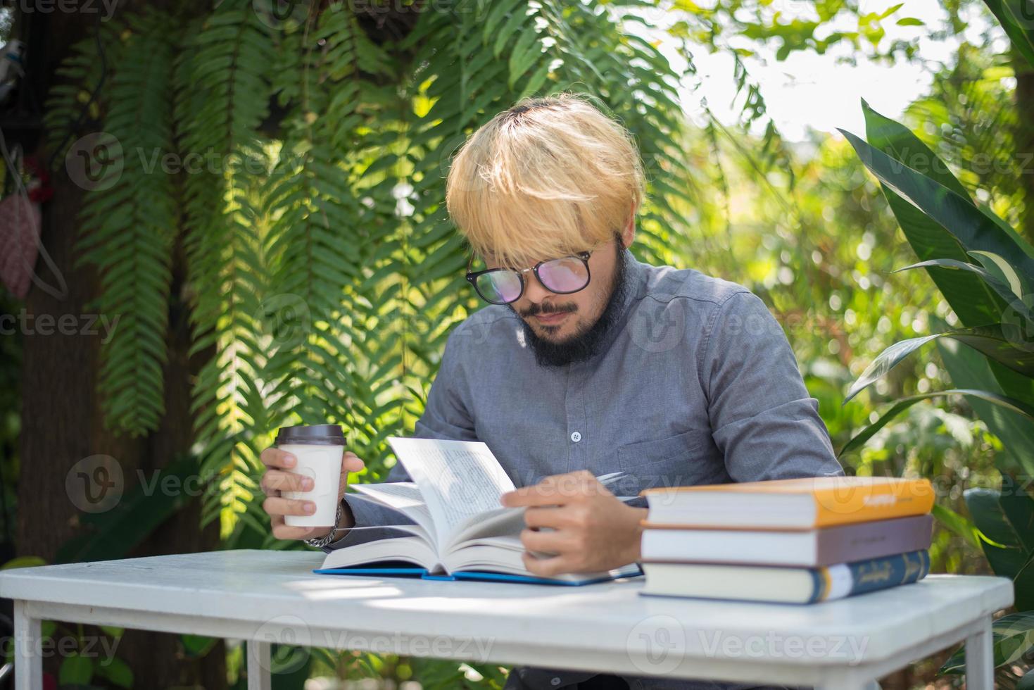 Hombre joven barba hipster tomando café mientras lee libros en el jardín de su casa con la naturaleza foto