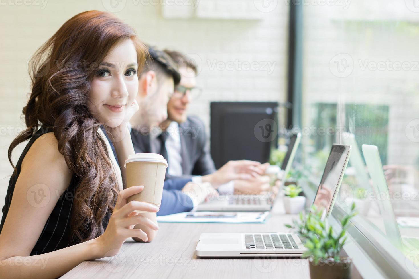 La empresaria tomando café mirando su computadora portátil mientras sus compañeros de trabajo interactúan en el fondo foto