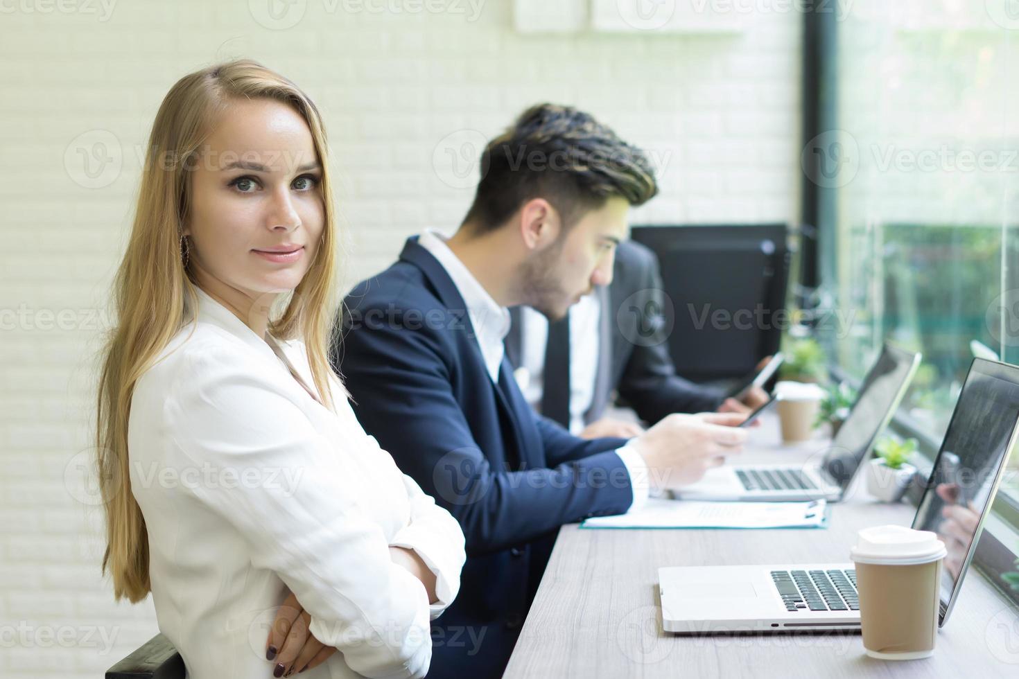 La empresaria usando la computadora portátil para trabajar mientras sus compañeros de trabajo interactúan en el fondo foto