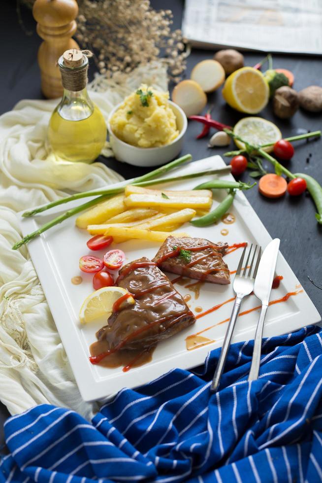 Bistec a la parrilla y papas fritas, con utensilios en un plato blanco con verduras en la mesa de madera oscura. foto