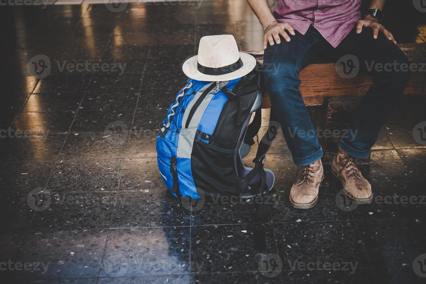 Hombre joven inconformista sentado en un banco de madera con mochila en la estación de tren foto