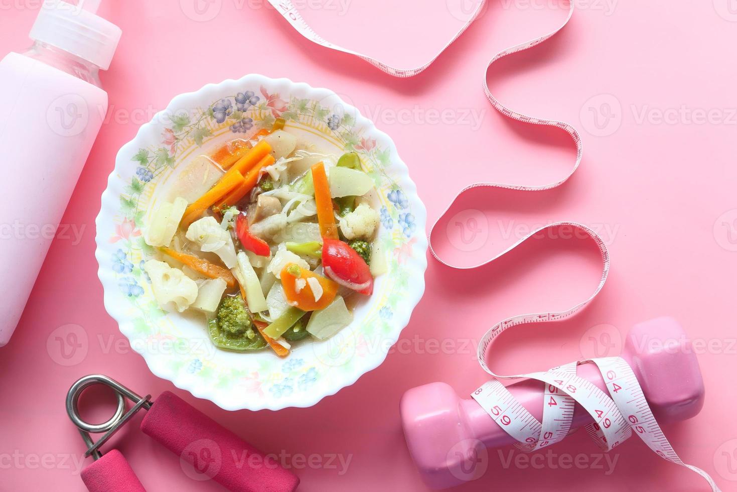 Fitness concept with dumbbell, fresh vegetables and measuring tape on pink background photo