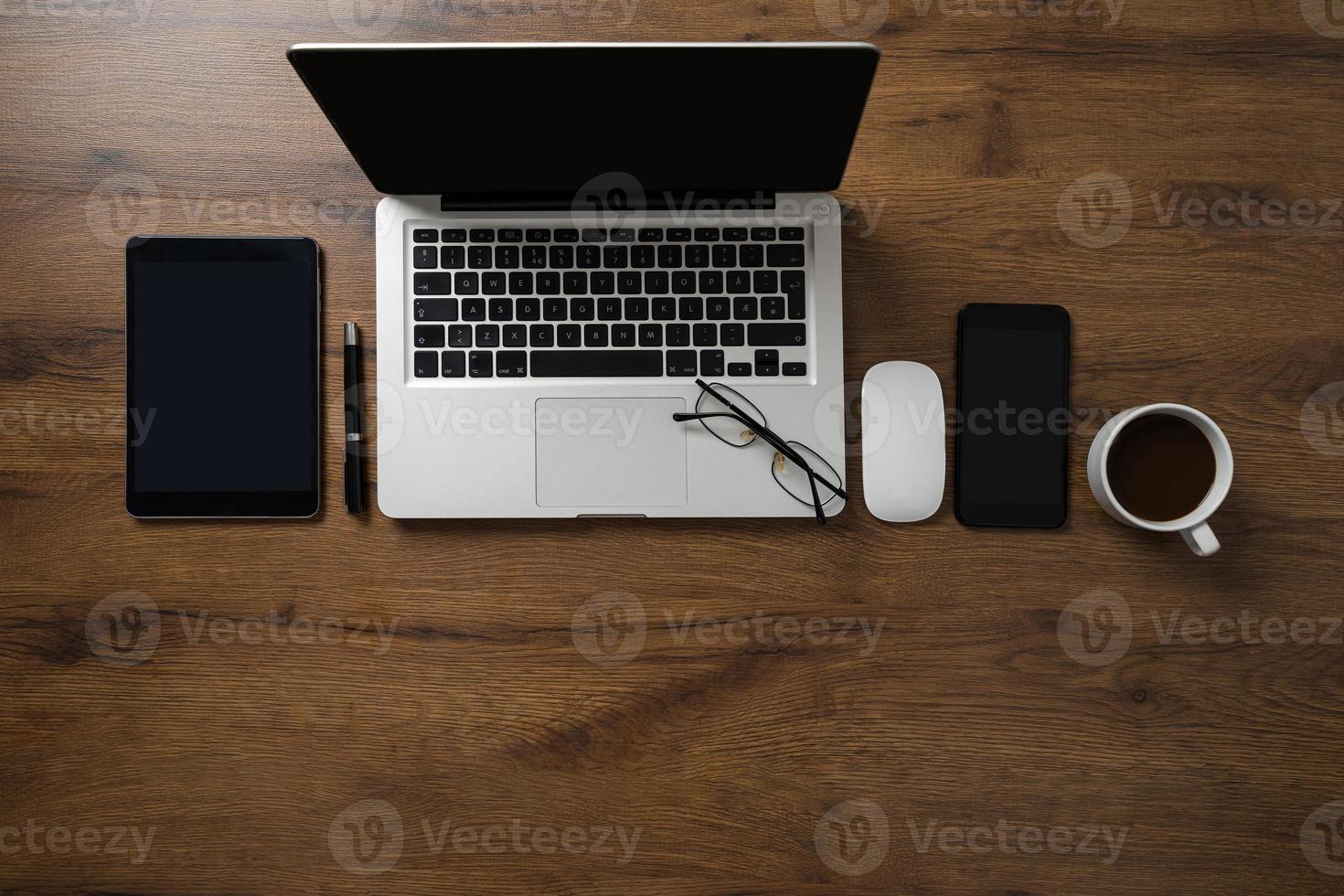 Vista superior del espacio de trabajo con computadora portátil, teléfono inteligente, tableta, taza de café, vasos y bolígrafo en la mesa de madera foto