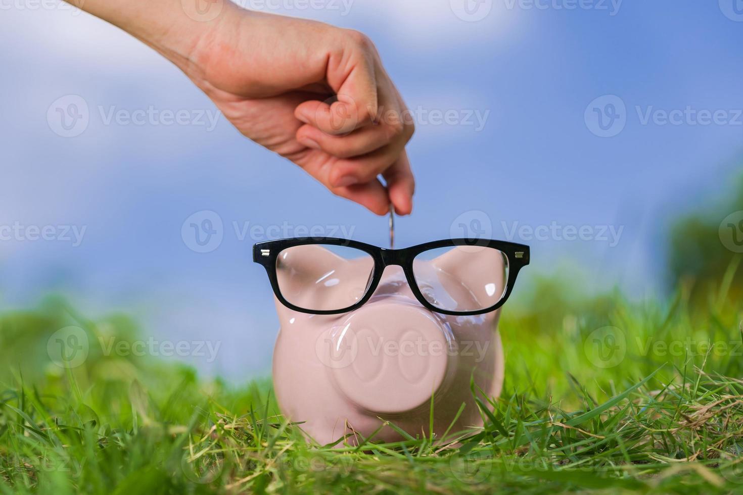 Pink piggy bank with glasses on grass and hand putting in a coin photo