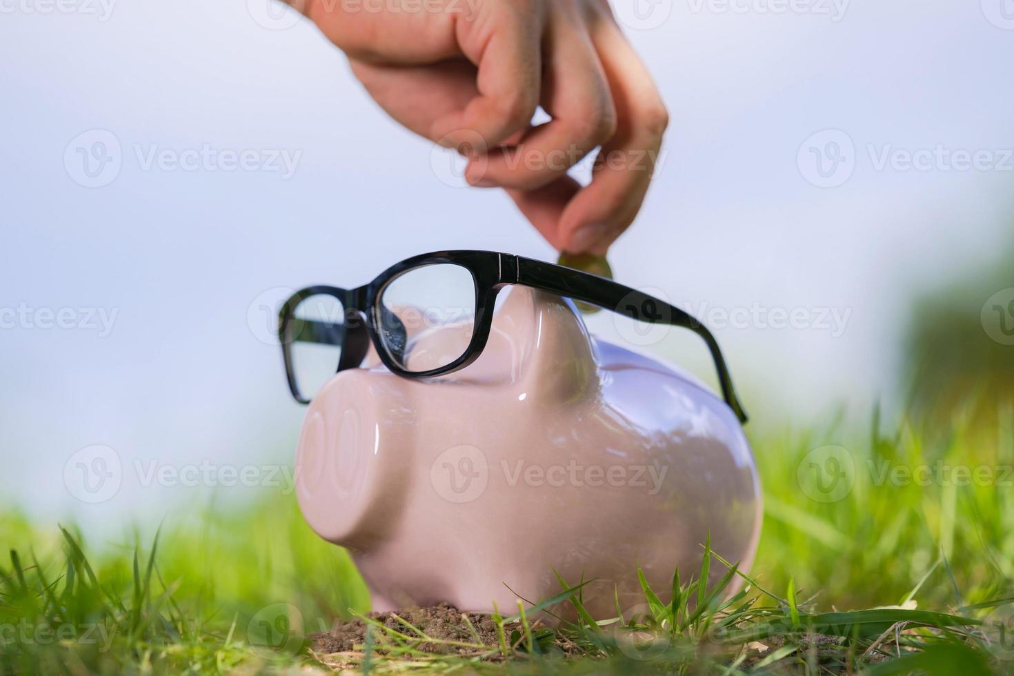 Pink piggy bank with glasses on grass and hand putting in a coin photo