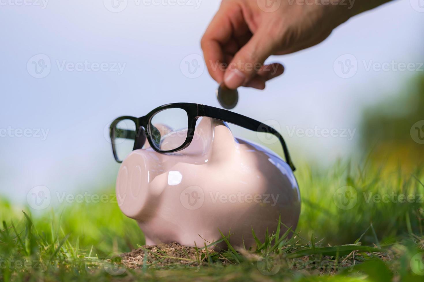 Pink piggy bank with glasses on grass and hand putting in a coin photo