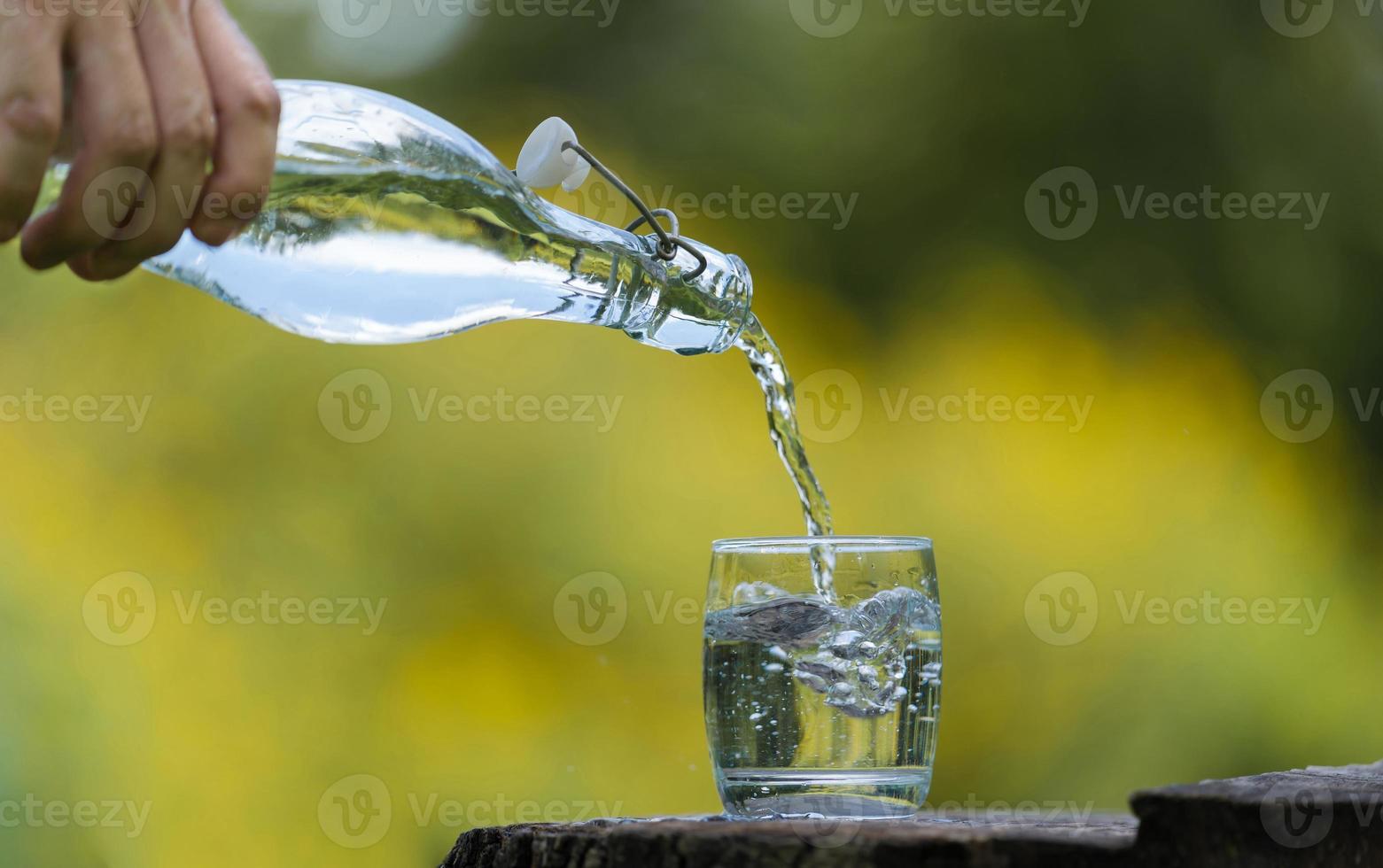 Mano vertiendo agua potable de botella en vaso con fondo natural foto