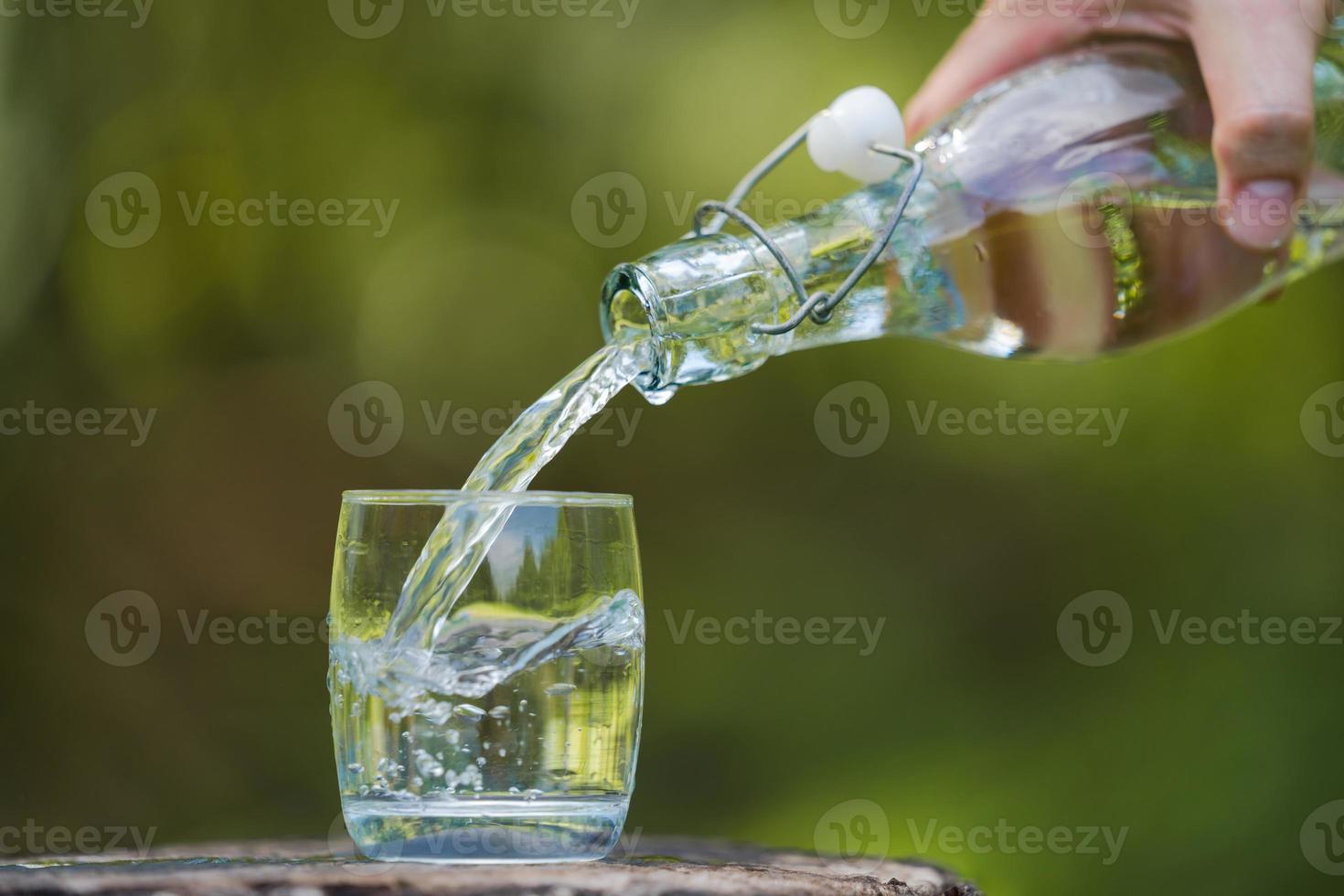 Mano vertiendo agua potable de botella en vaso con fondo natural foto