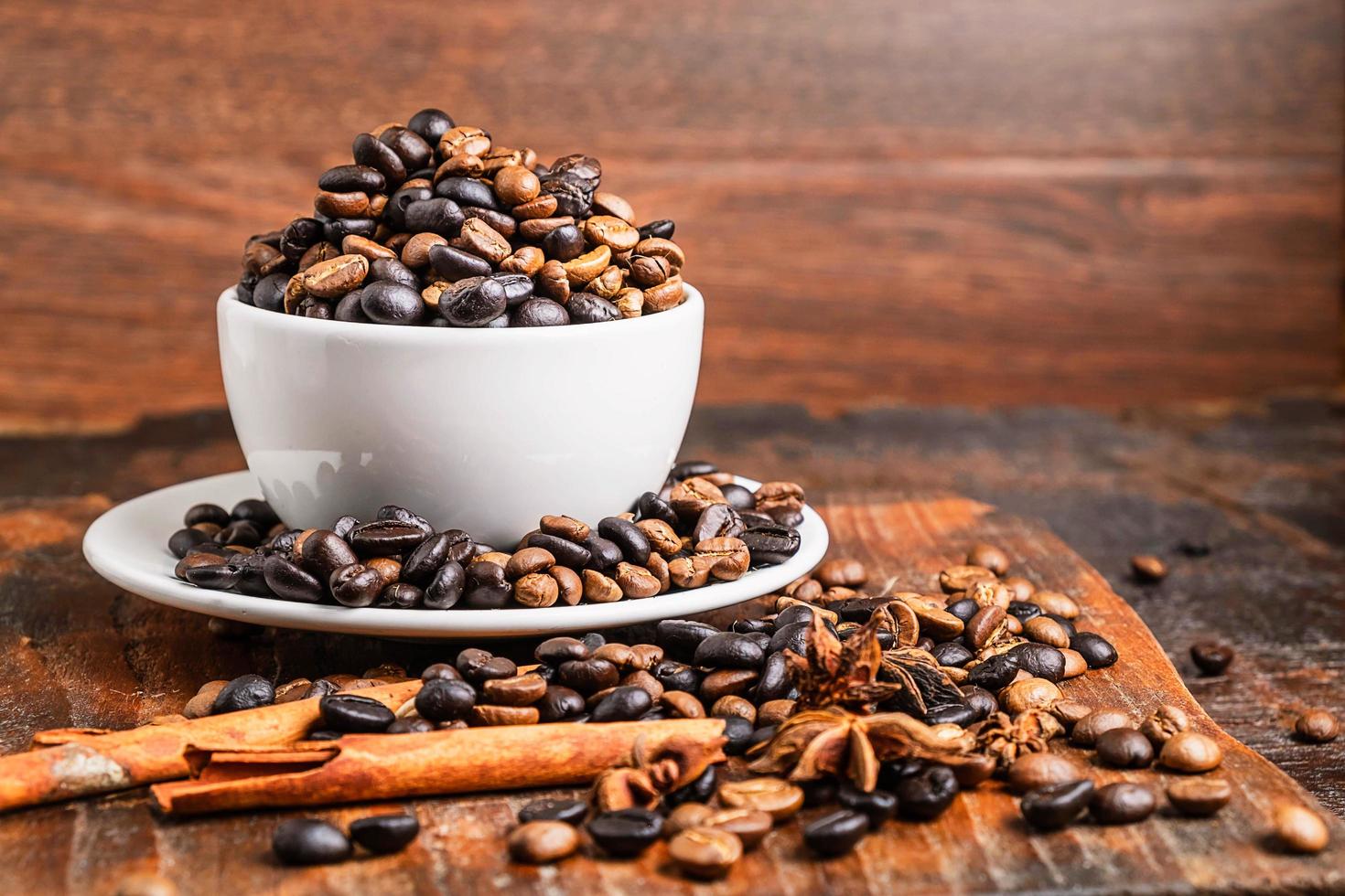 Granos de café en una taza blanca con platillo y ramitas de canela sobre una tabla para cortar madera sobre una mesa de madera oscura. foto