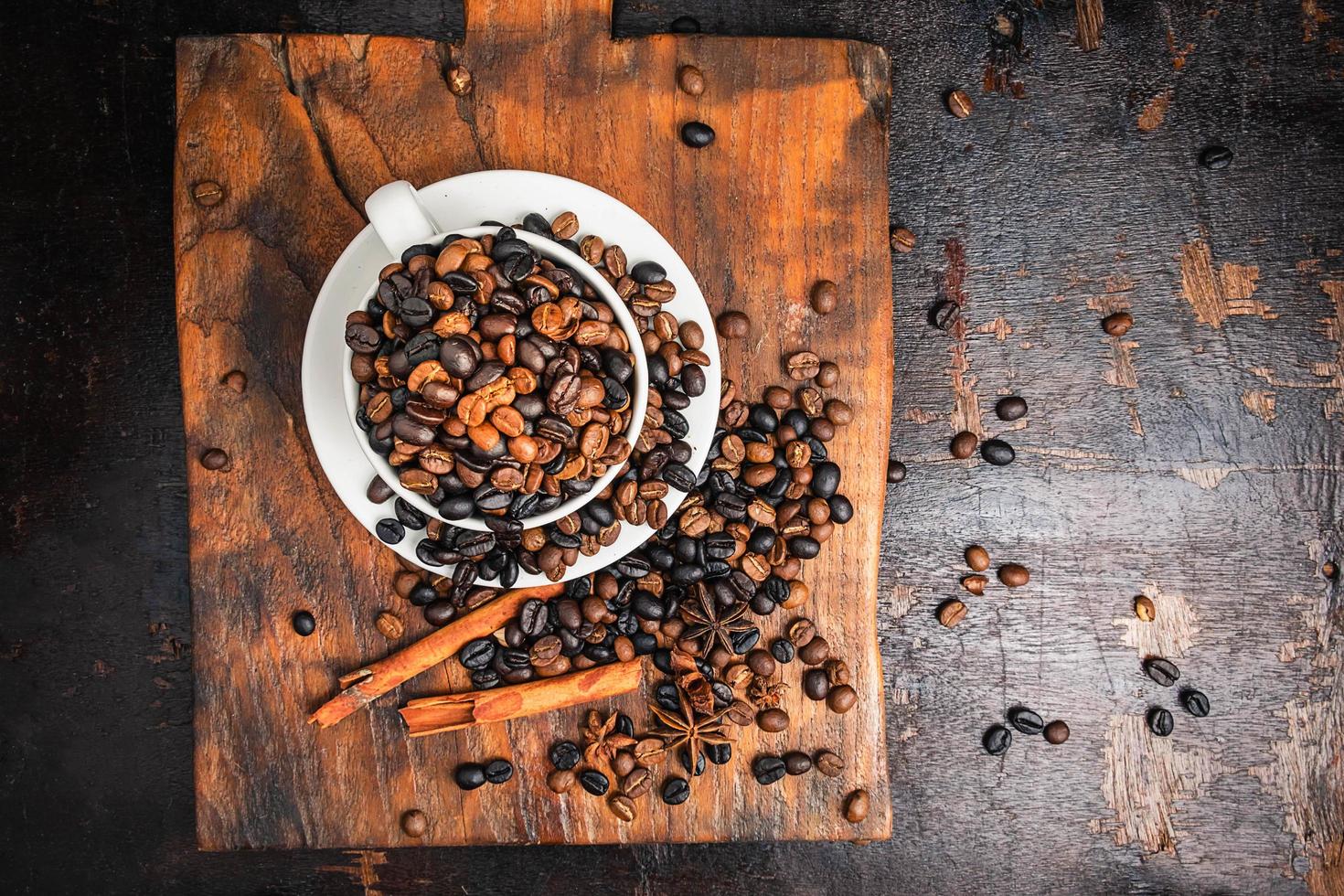 Granos de café en una taza blanca con platillo y ramitas de canela sobre una tabla para cortar madera sobre una mesa de madera oscura. foto