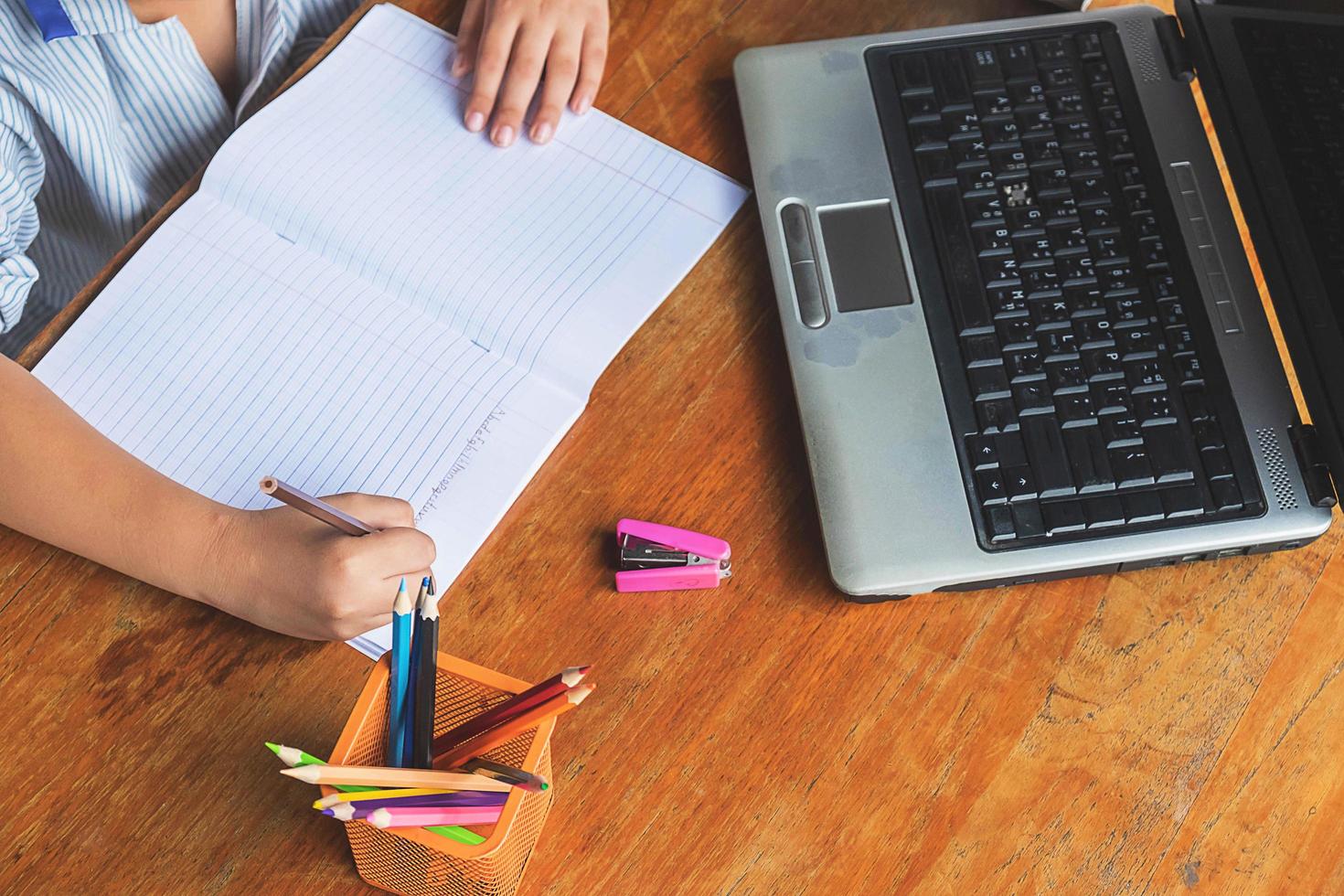 Niño haciendo los deberes con el cuaderno, la computadora portátil, la grapadora y una taza de lápices en un escritorio de madera foto