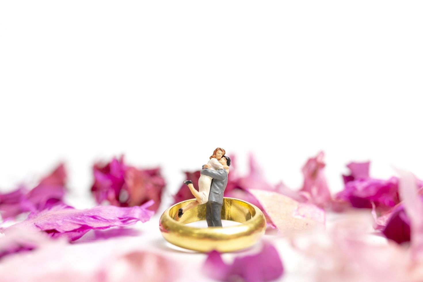 Pareja en miniatura abrazándose con un anillo de bodas y pétalos de rosa aislado sobre un fondo blanco. foto
