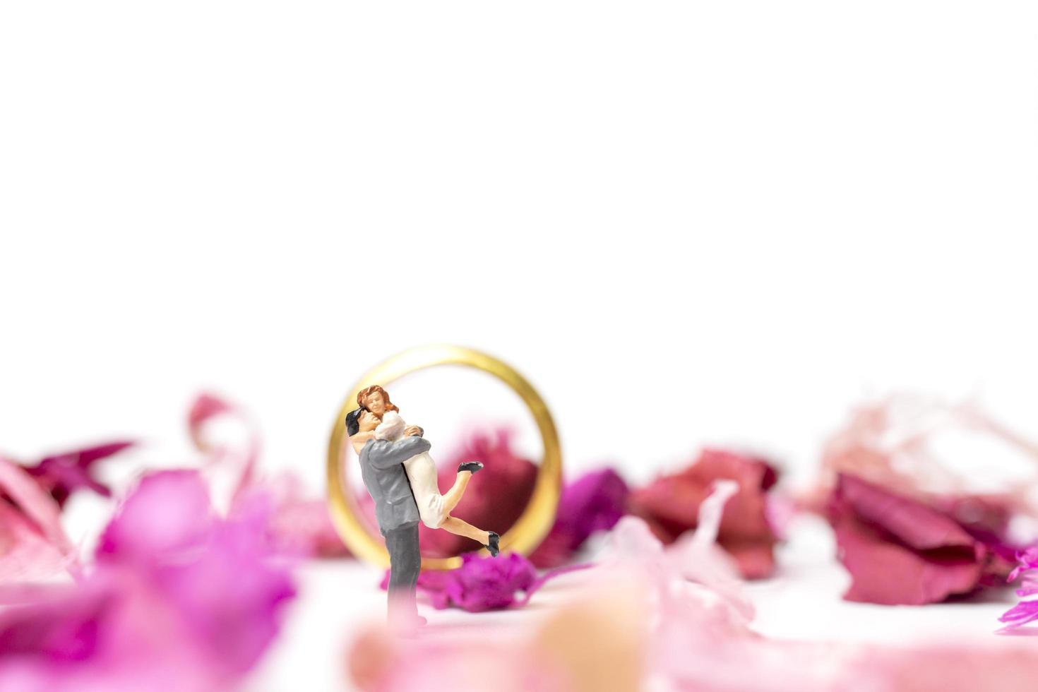 Pareja en miniatura abrazándose con un anillo de bodas y pétalos de rosa aislado sobre un fondo blanco. foto