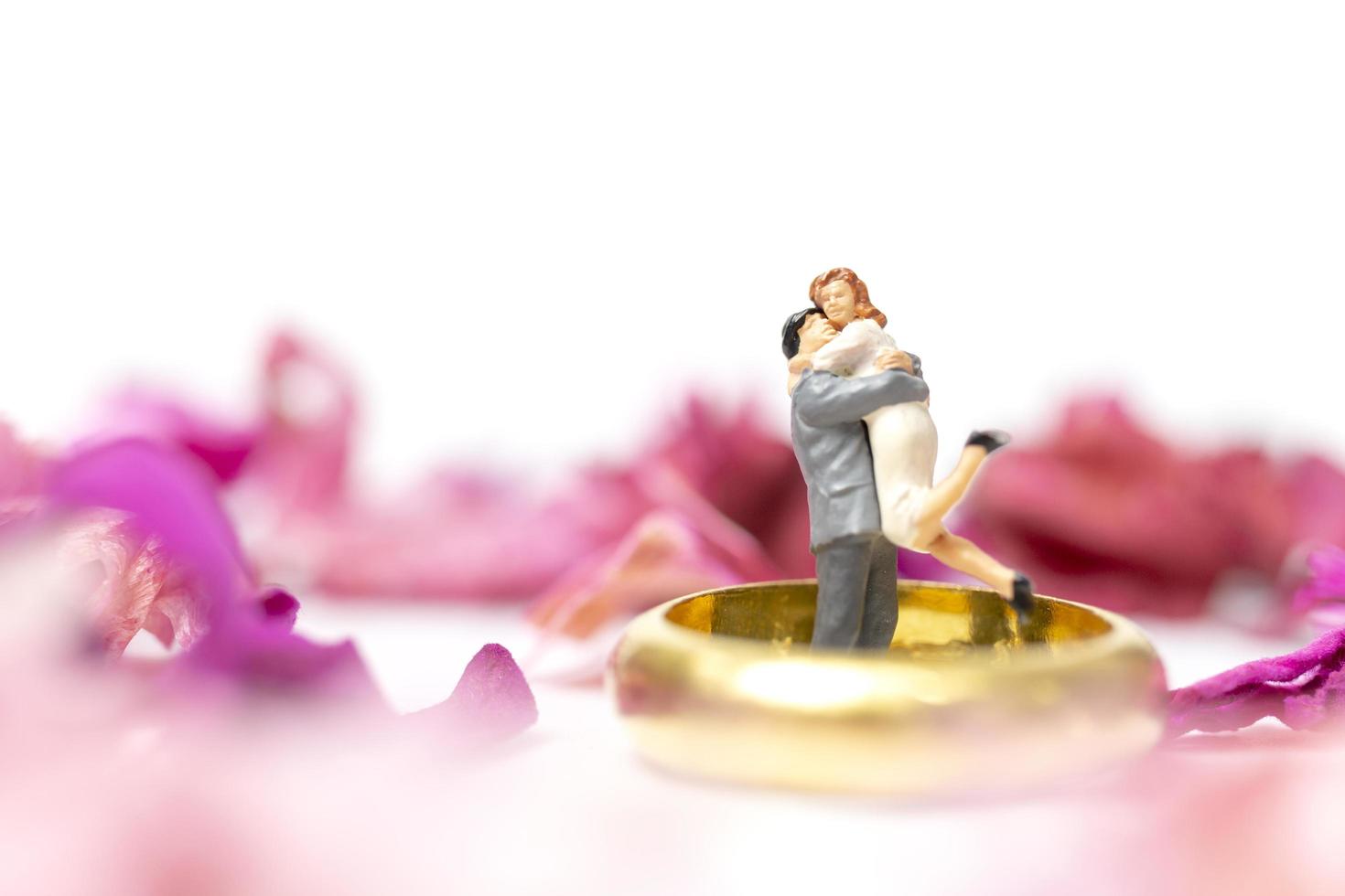 Pareja en miniatura abrazándose con un anillo de bodas y pétalos de rosa aislado sobre un fondo blanco. foto
