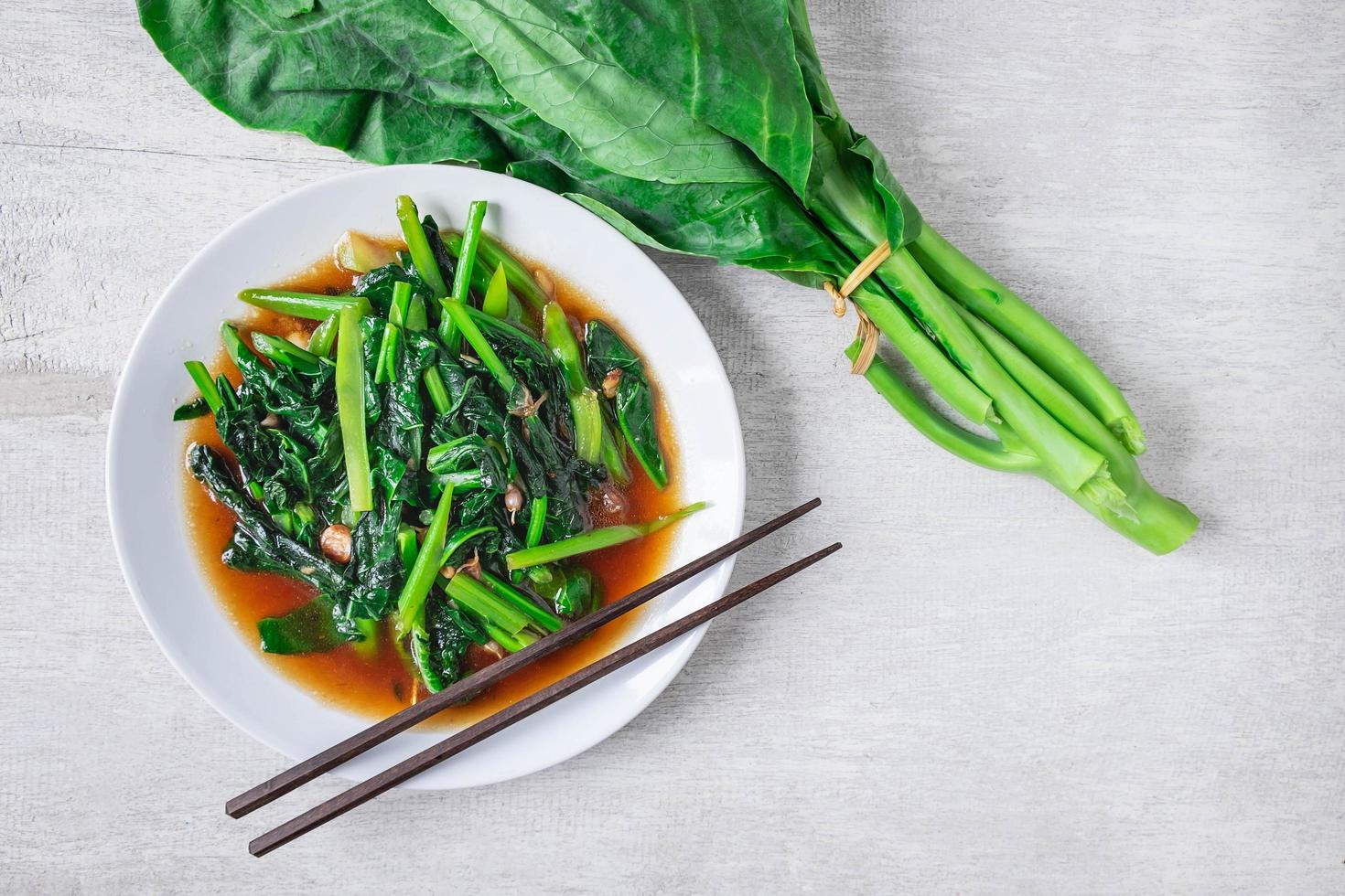 Fried Chinese kale with oyster sauce with chopsticks next to fresh Chinese kale on a white plate on wooden table photo