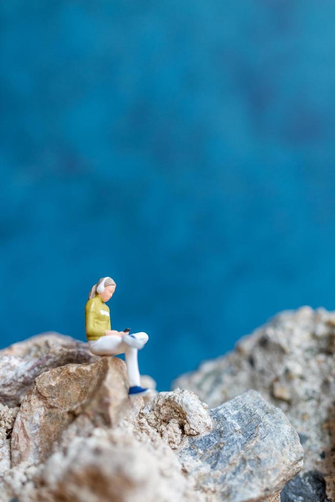 Mujer en miniatura usando audífonos y escuchando música en un teléfono inteligente sentado en la roca foto