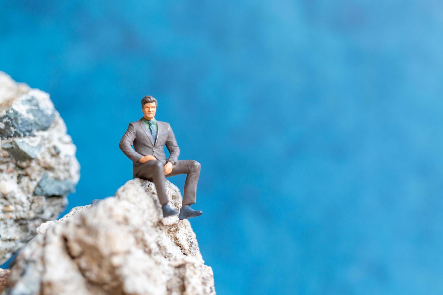 Empresario en miniatura sentado sobre una roca con un fondo azul. foto