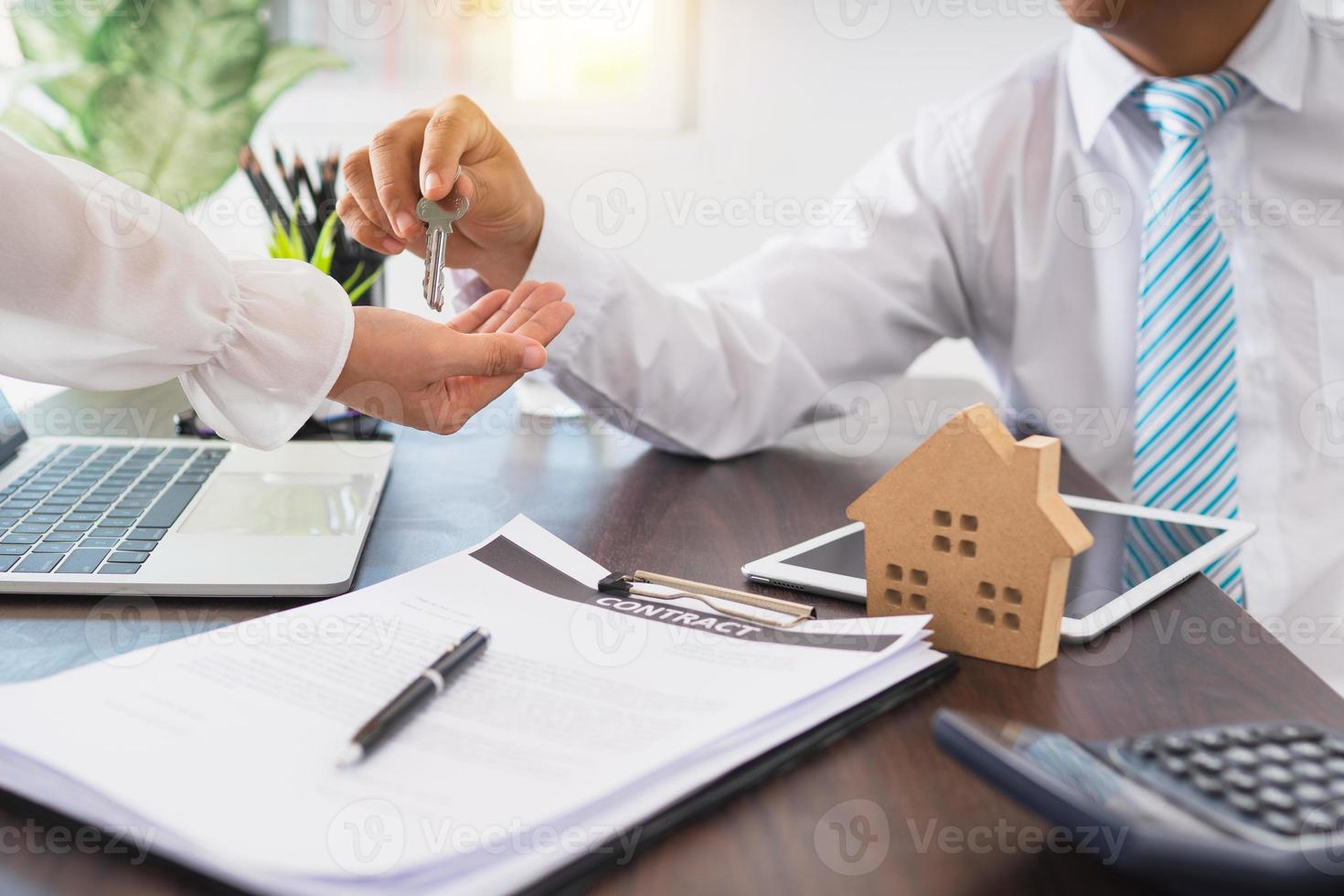 Businessman hands a key to a person next to laptop, contract, and model house photo