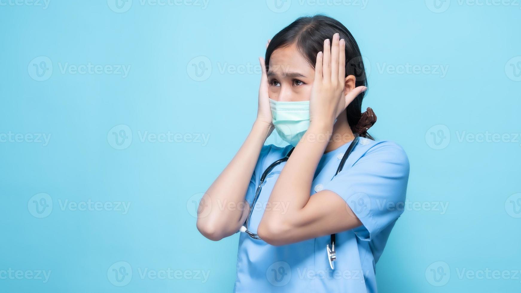 Mujer asiática en uniforme médico azul con una mascarilla y un estetoscopio gesticulando con las manos contra el costado de su cabeza contra el fondo azul. foto