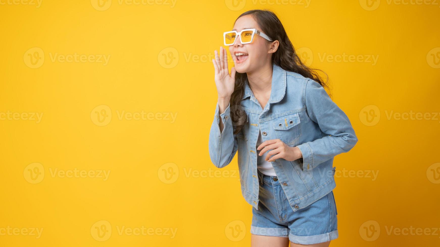 Mujer asiática sonriendo y gesticulando con la mano abierta junto a la boca sobre fondo amarillo foto