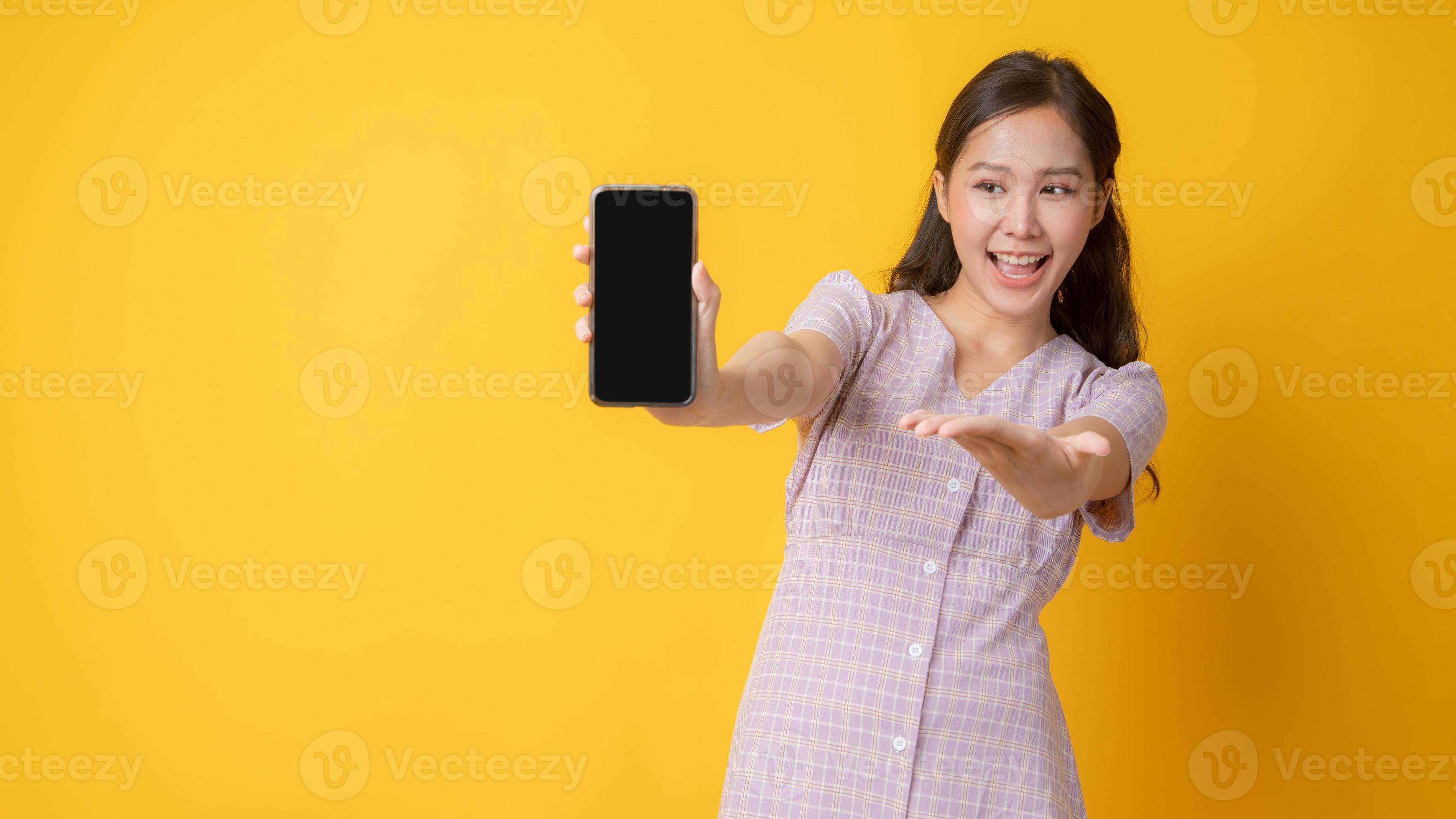 Asian woman gesturing toward a blank black cell phone on yellow background photo