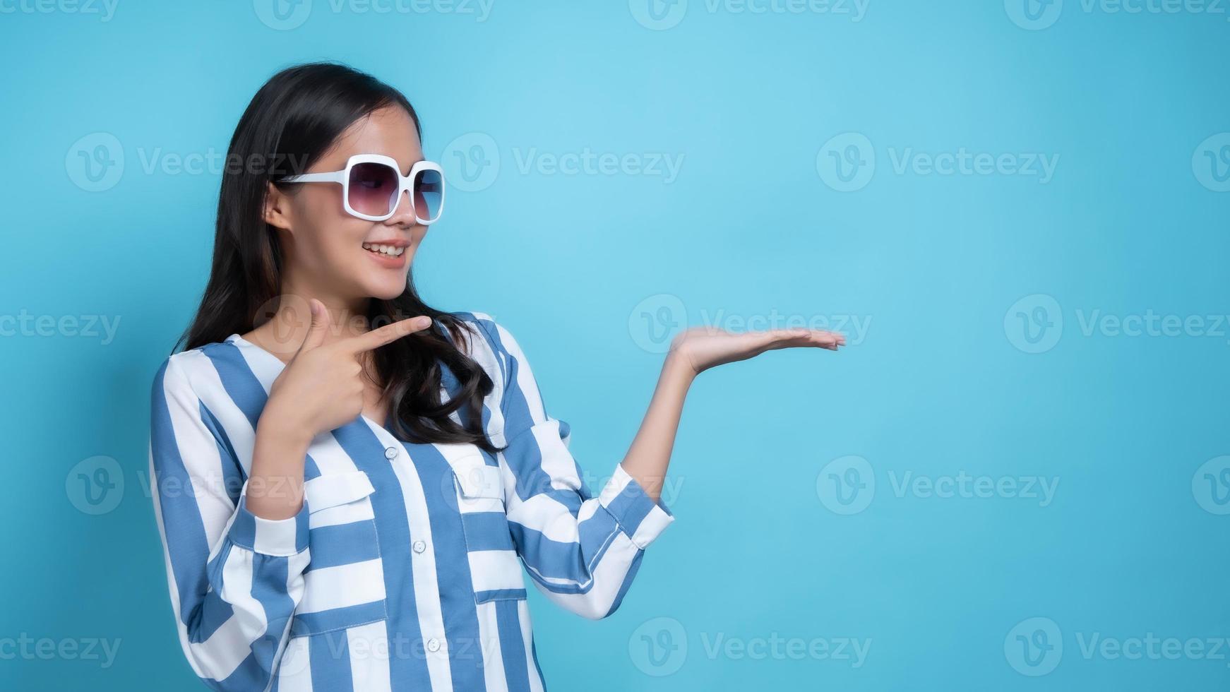 Mujer asiática con gafas de sol blancas apuntando hacia el espacio de la copia sobre fondo azul. foto