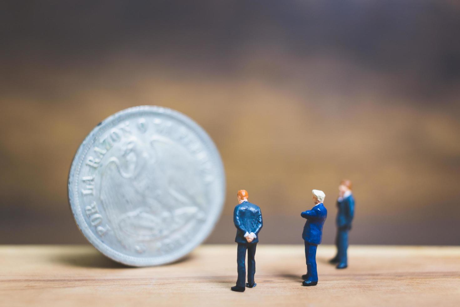 Miniature businessmen standing near a coin with a wooden background, business concept photo