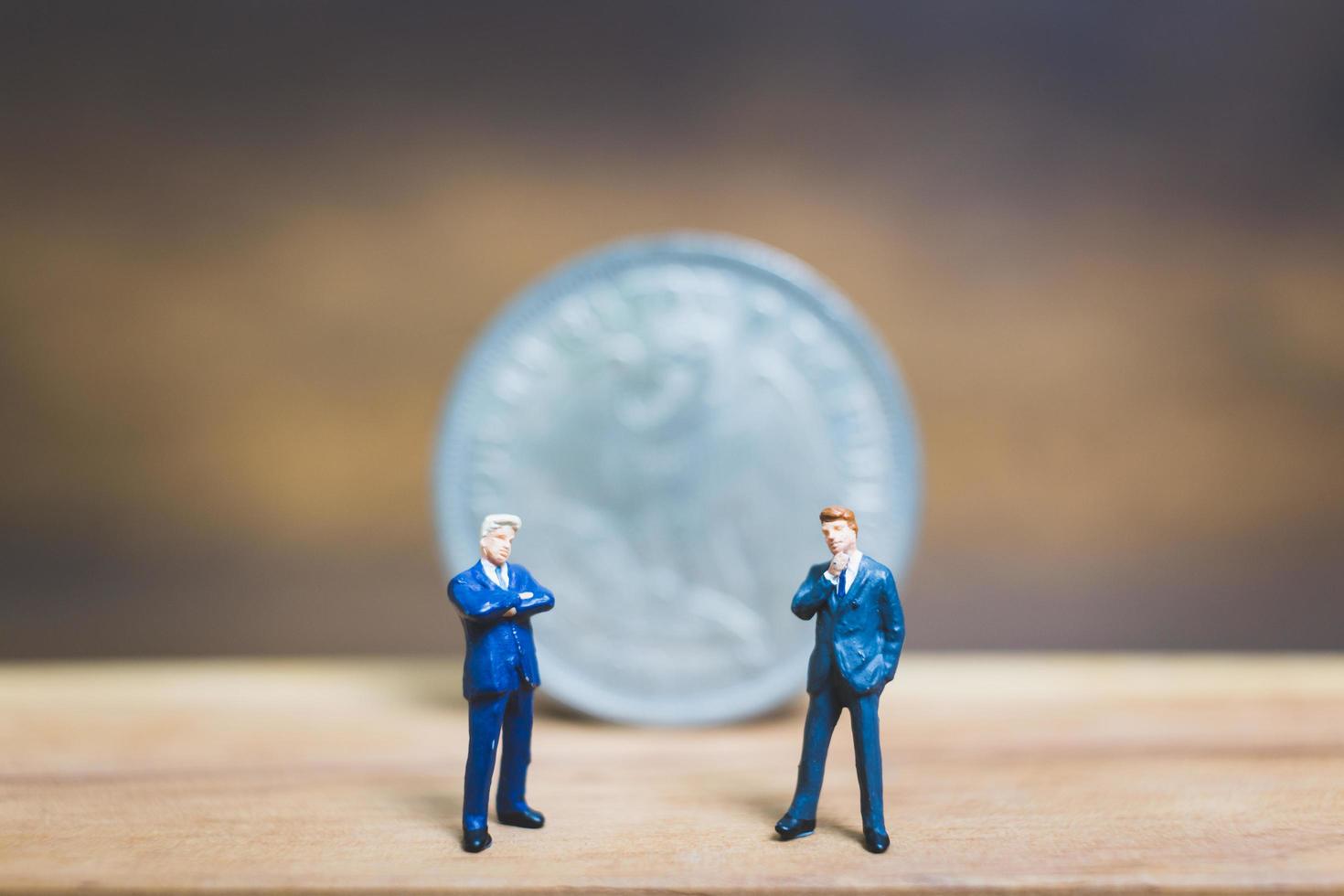 Miniature businessmen standing near a coin with a wooden background, business concept photo