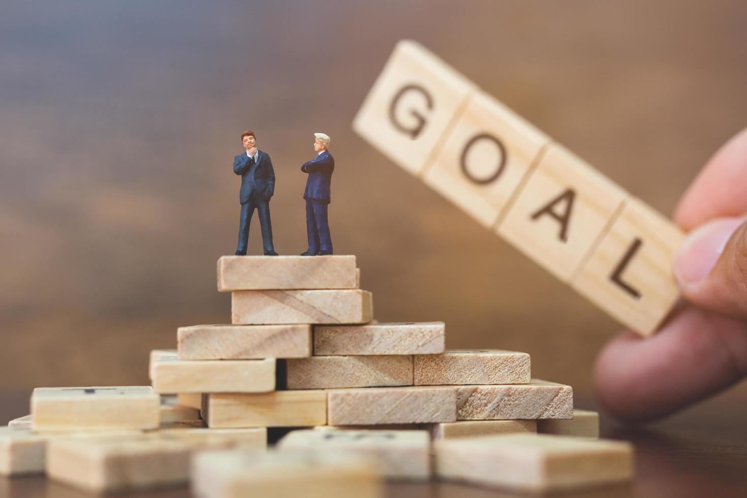 Miniature businessmen standing on wooden blocks with a hand holding the word Goal, business career growth concept photo