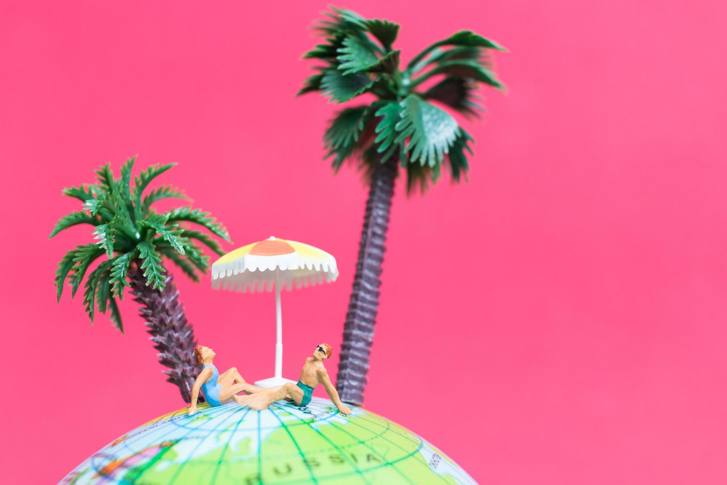 Miniature people wearing swimsuits relaxing on a globe with a pink background, honeymoon concept photo