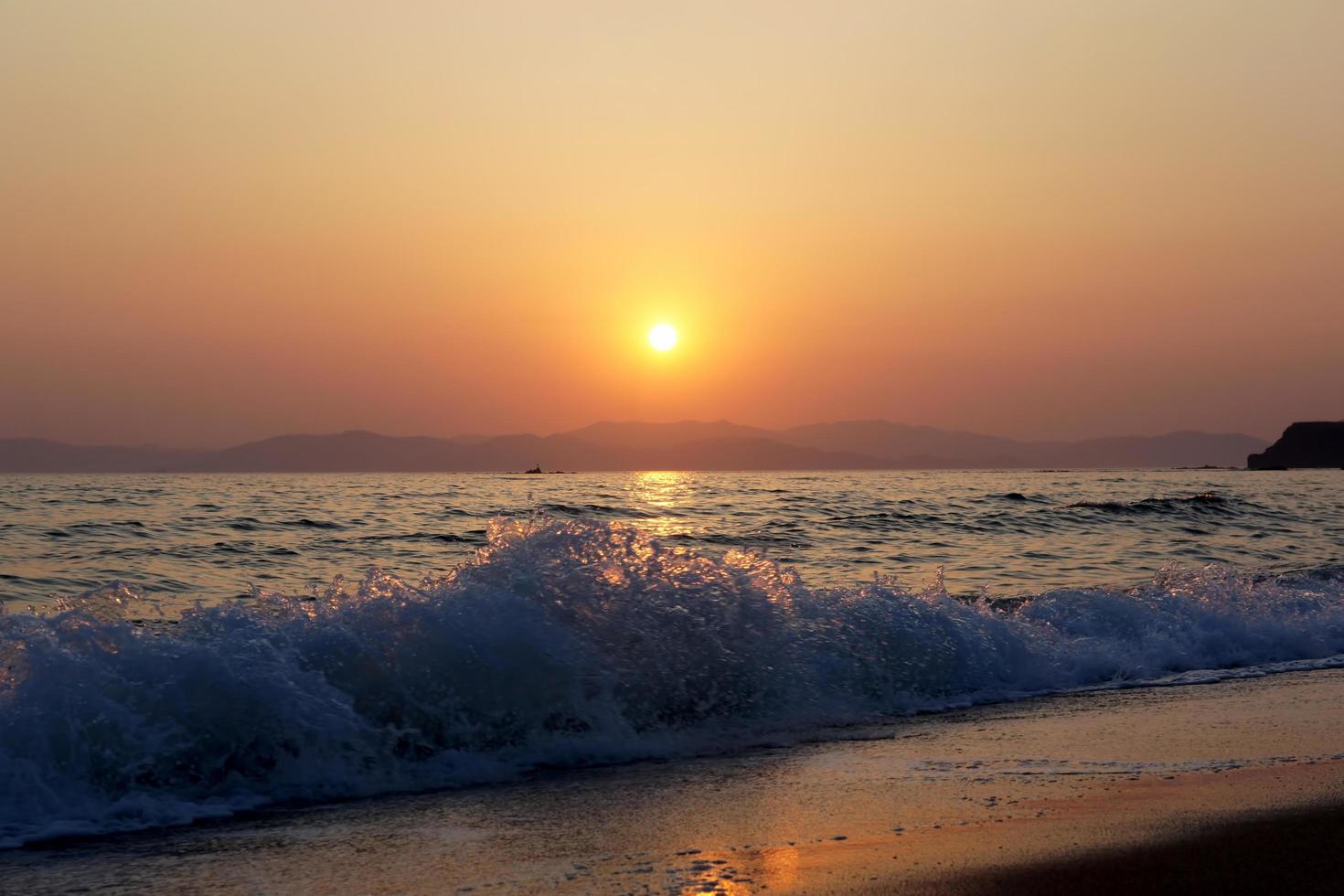 Olas rompiendo en una playa con atardecer nublado naranja sobre las montañas foto