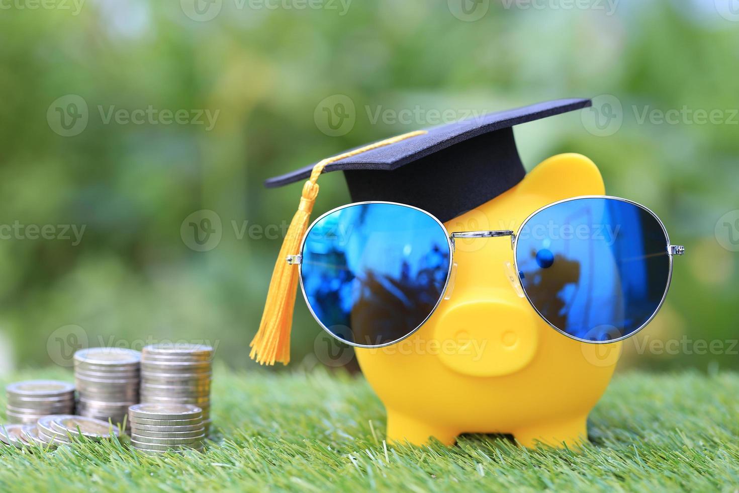 Sombrero de graduación en una alcancía dorada con gafas de sol y una pila de monedas sobre un fondo verde natural, ahorrando dinero para el concepto de educación foto