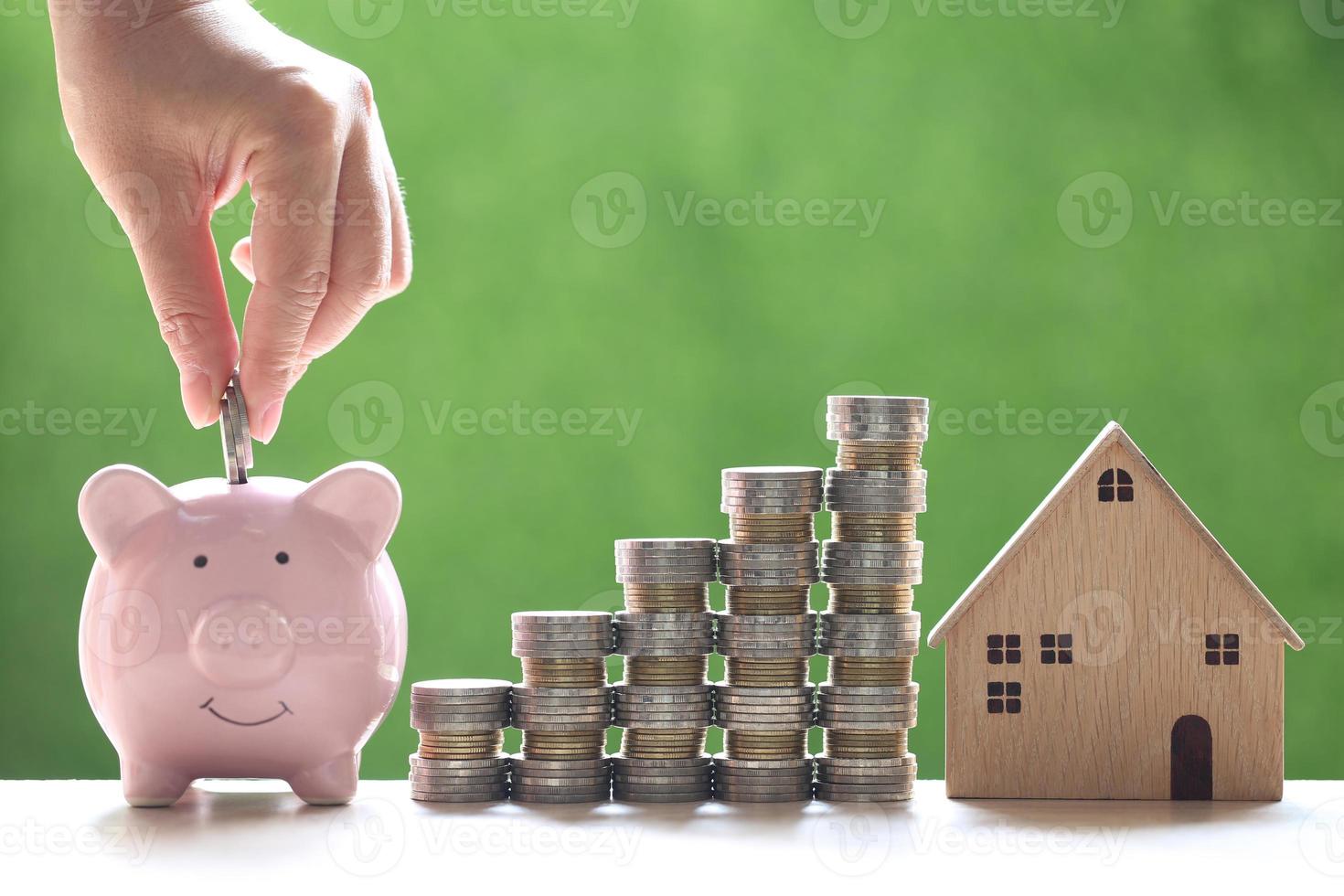 Casa modelo con montones de monedas junto a una mano poniendo dinero en una alcancía sobre un fondo verde natural, ahorrando dinero para la preparación del futuro y el concepto de inversión foto