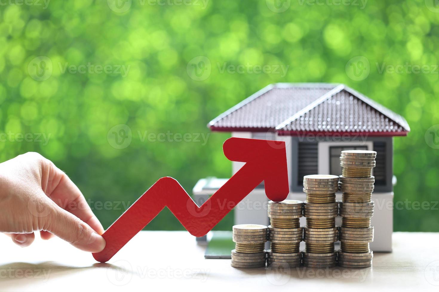 A woman's hand holding a red arrow on a stack of coins money with a model house on a natural green background, business investment and real estate concept photo