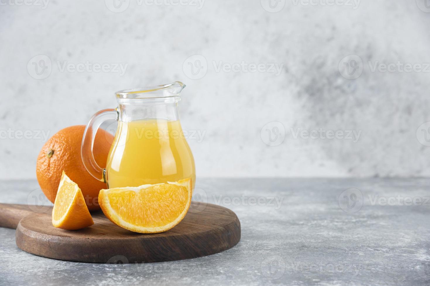 A glass pitcher of fresh juice with whole and sliced orange fruit placed on a wooden board photo